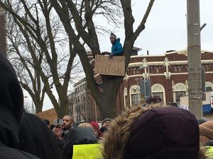 Augustana first year Mikaylo Kelly gets on top of a tree to show off his sign amidst the protest on Saturday, Feb. 4.