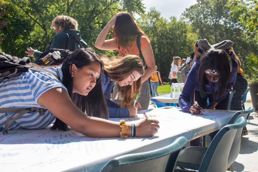 Augustana seniors Samantha Dinglasan (left), Vanessa Blankson (right), and junior Vlada Botsul (center) write their pledges to be inclusive on a poster at the Inclusive Augie Campaign Kickoff, Augustana College lower quad, September 6th, 2019.