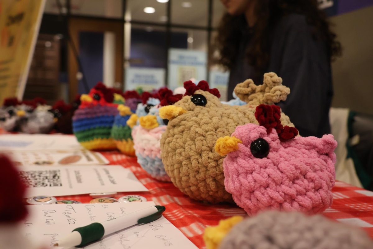 Handmade crochet chickens in various colors are displayed on a table alongside sign-up sheets during a fundraising event. The collaboration between Soan Society and Yarn Club aims to support the River Bend Food Bank through the sale of the crafted items.
